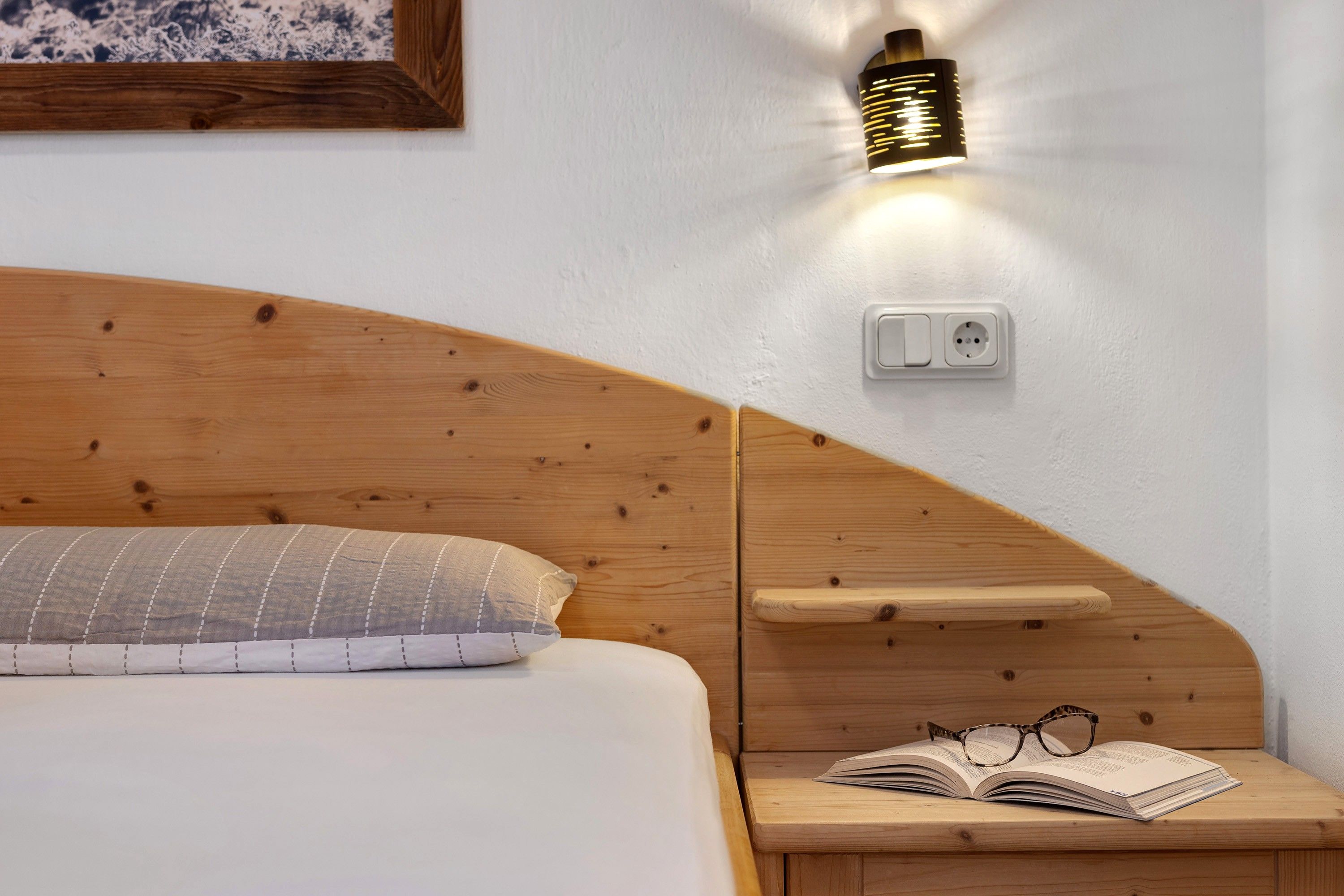 A bedroom with a wooden headboard, a bedside table with a book and glasses, a wall-mounted light fixture, and a power outlet.