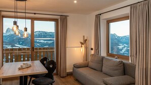 Une pièce chaleureuse avec une table à manger en bois, deux chaises, un canapé et de grandes fenêtres donnant sur les montagnes.