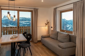 Une pièce chaleureuse avec une table à manger en bois, deux chaises, un canapé et de grandes fenêtres donnant sur les montagnes.