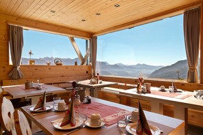 A wooden interior with a dining table set for two, a view of mountains through large windows, and a wooden ceiling with recessed lighting.