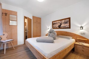 A bedroom with a wooden bed, a chair, a bedside table, a nightstand, and a wall-mounted picture.