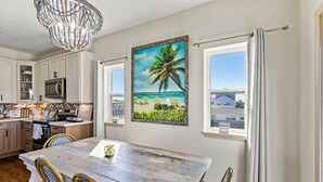 A kitchen with white cabinets, a wooden table, wicker chairs, and a painting of a beach scene. - Relaxing Beach Escape w/ Private Decks Walk2Beach (Galveston)
