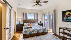 A bedroom with a bed, a ceiling fan, a window, a nightstand, a lamp, a rug, and a door. - Relaxing Beach Escape w/ Private Decks Walk2Beach (Galveston)