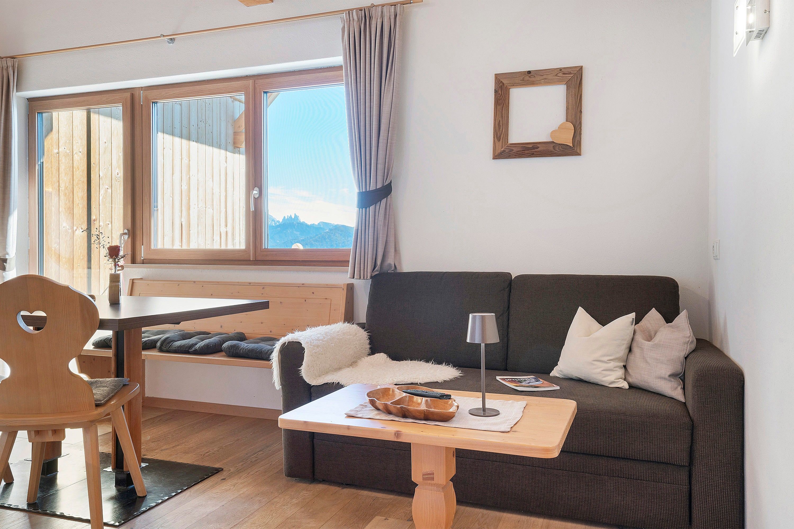 Una sala acogedora con una mesa de madera, un sofá con cojines, una mesita con una lámpara y una ventana con cortinas.