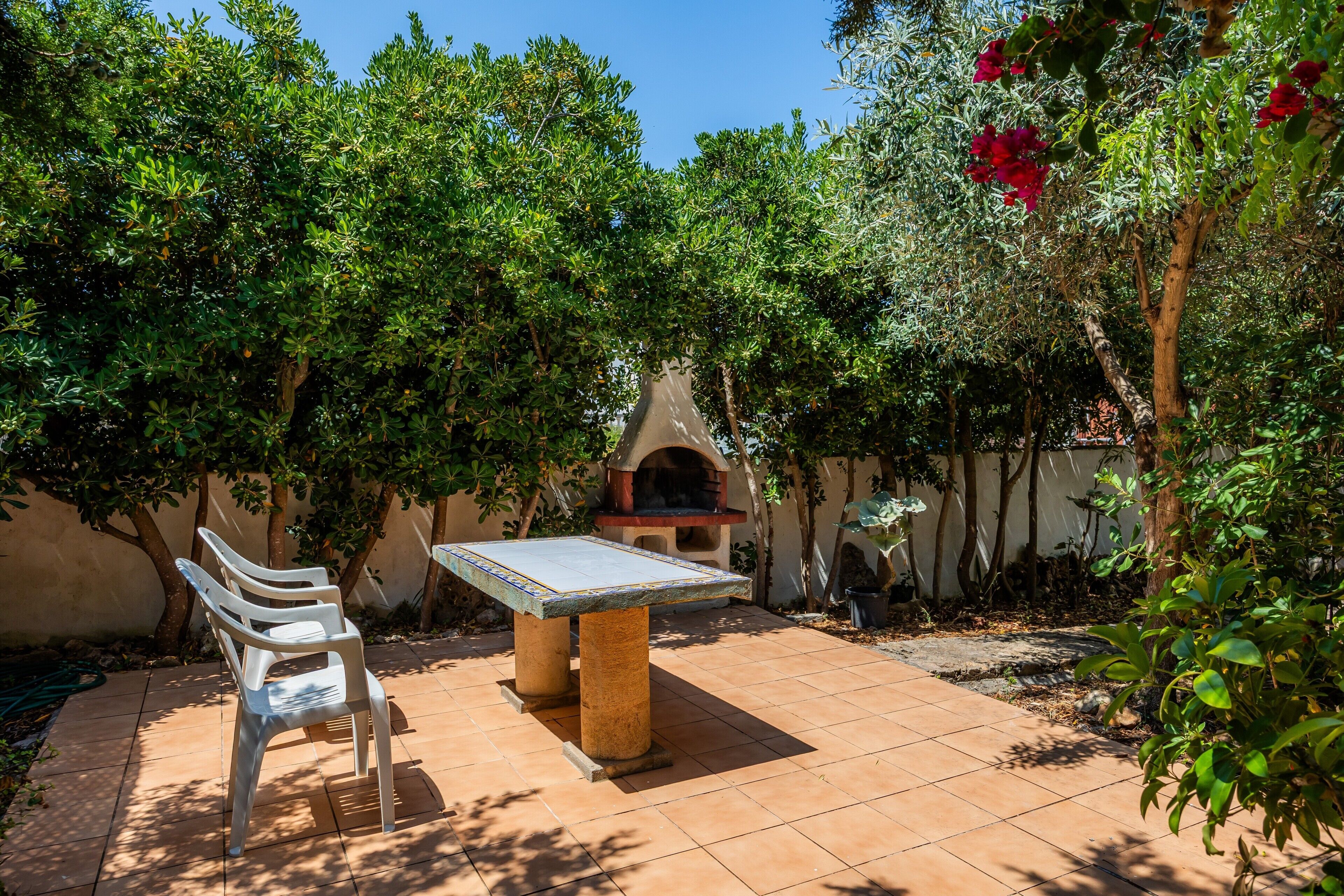Um pátio externo com mesa e cadeiras, cercado por vegetação exuberante e um forno de tijolos.