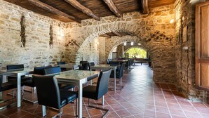 Un coin repas rustique aux murs de pierre, avec des tables et des chaises, un plafond en bois et une vue sur l’extérieur à travers une fenêtre en arc de cercle.