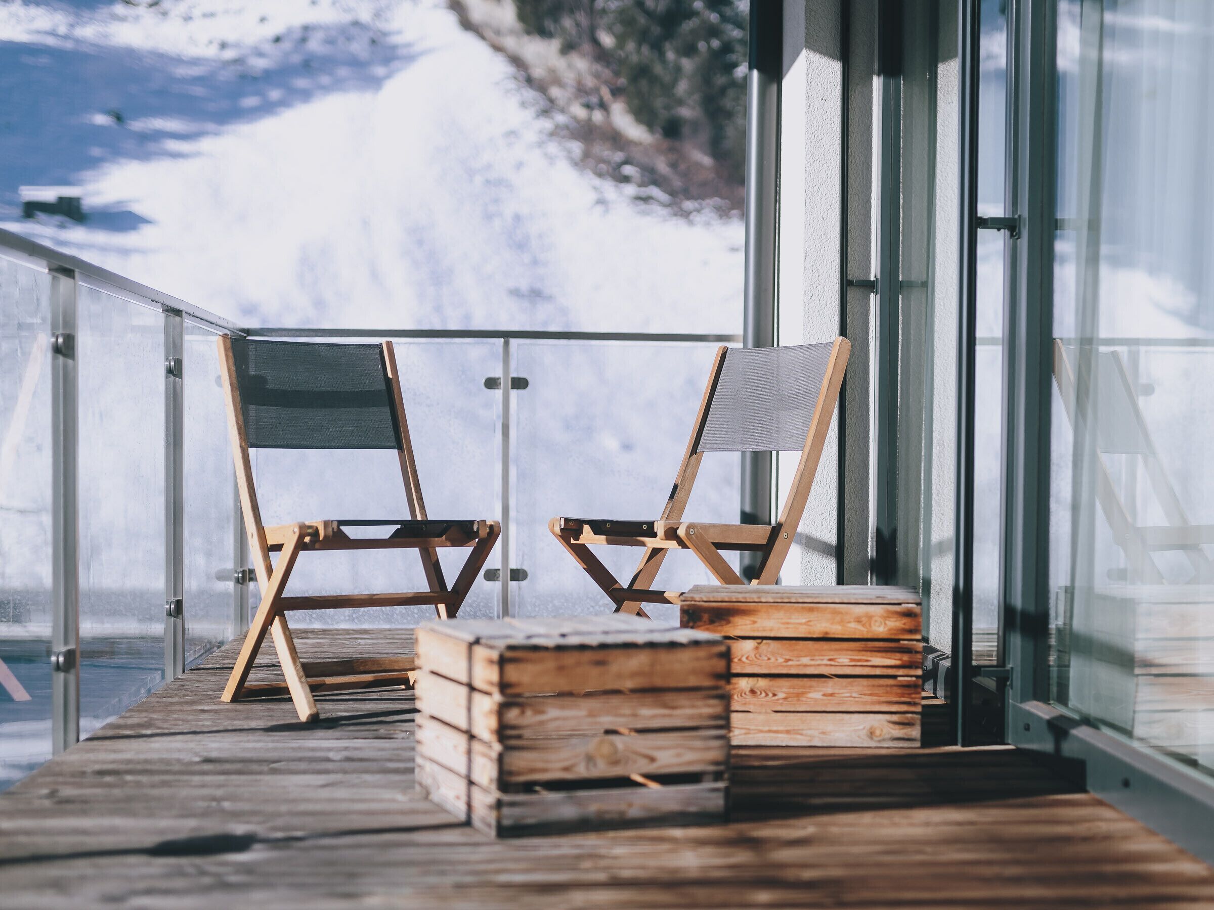 Two wooden deck chairs with canvas seats and backs, stacked wooden crates, and a glass railing overlooking a snowy landscape.
