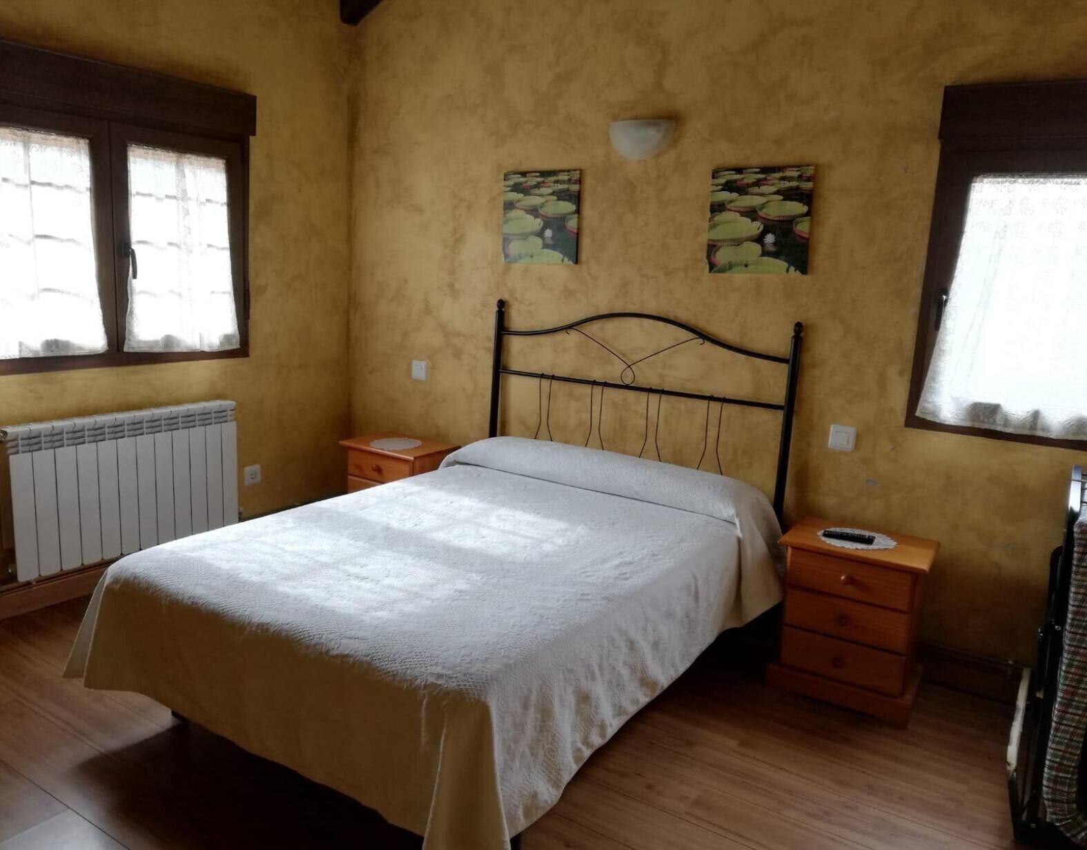 A bedroom with a bed, two bedside tables, a radiator, and two windows with curtains.
