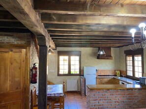 Uma cozinha rústica com vigas de madeira aparentes, painel de parede de tijolos, uma mesa de jantar com cadeiras e uma geladeira.