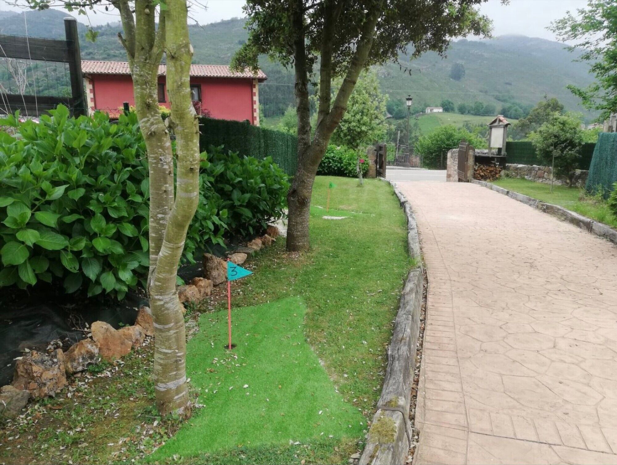 A paved pathway leading to a building with a red facade, surrounded by greenery and a hilly landscape.