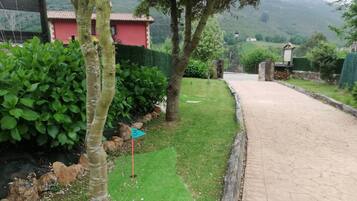 A paved pathway leading to a building with a red facade, surrounded by greenery and a hilly landscape.
