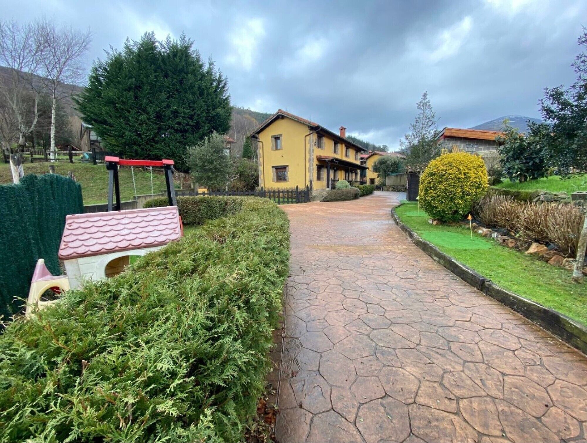 Un chemin pavé mène à un bâtiment jaune surmonté d’un toit rouge, entouré de verdure et agrémenté d’une petite niche à chien.