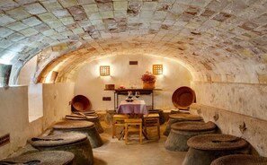 An arched stone room with a table and stools, a small shelf with wine glasses, and a potted plant.