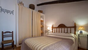 Ein Schlafzimmer mit einem großen Bett, einem Holzstuhl, einem Nachttisch mit Lampe und einem Regal mit Gegenständen.