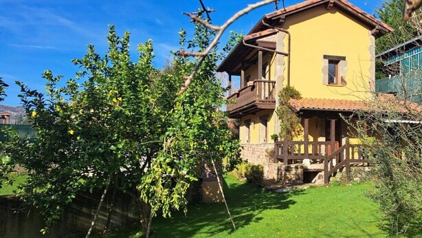 A two-story house with a balcony, surrounded by greenery and a stone wall.