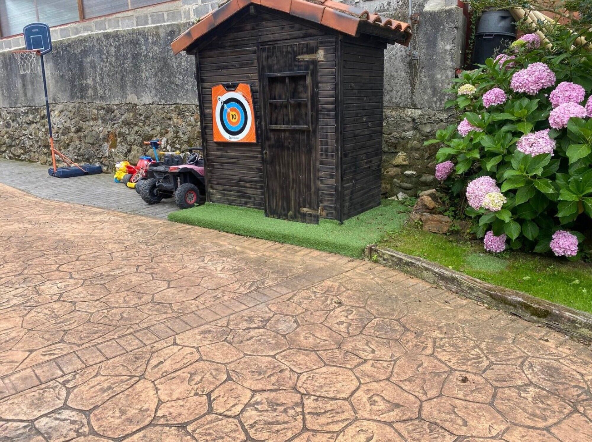 A stone-paved area with a small wooden structure, a target, and a pink flowering bush.
