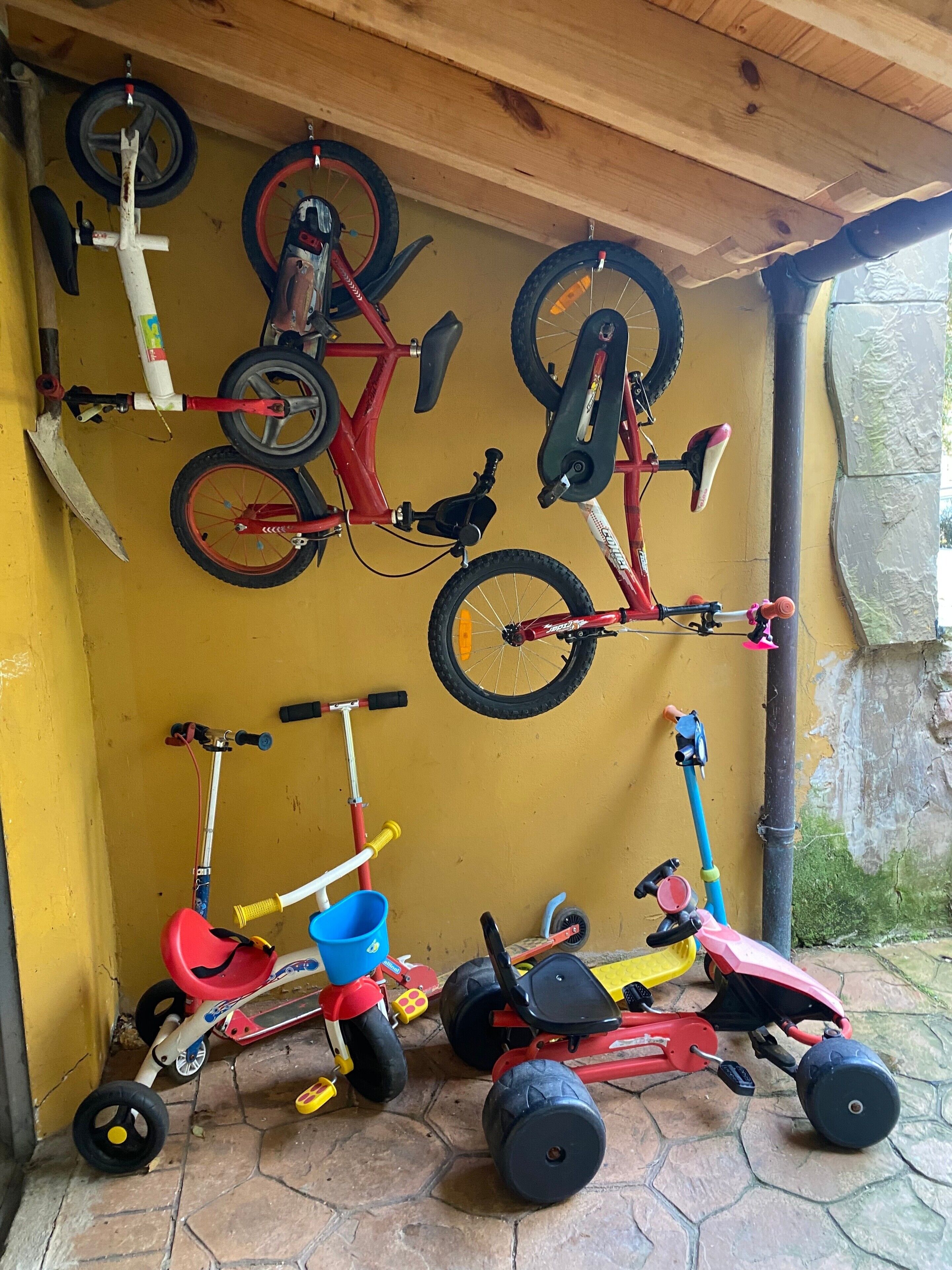 Een overdekte ruimte met fietsen en driewielers aan de muur, een scooter en een blauwe emmer op de vloer.
