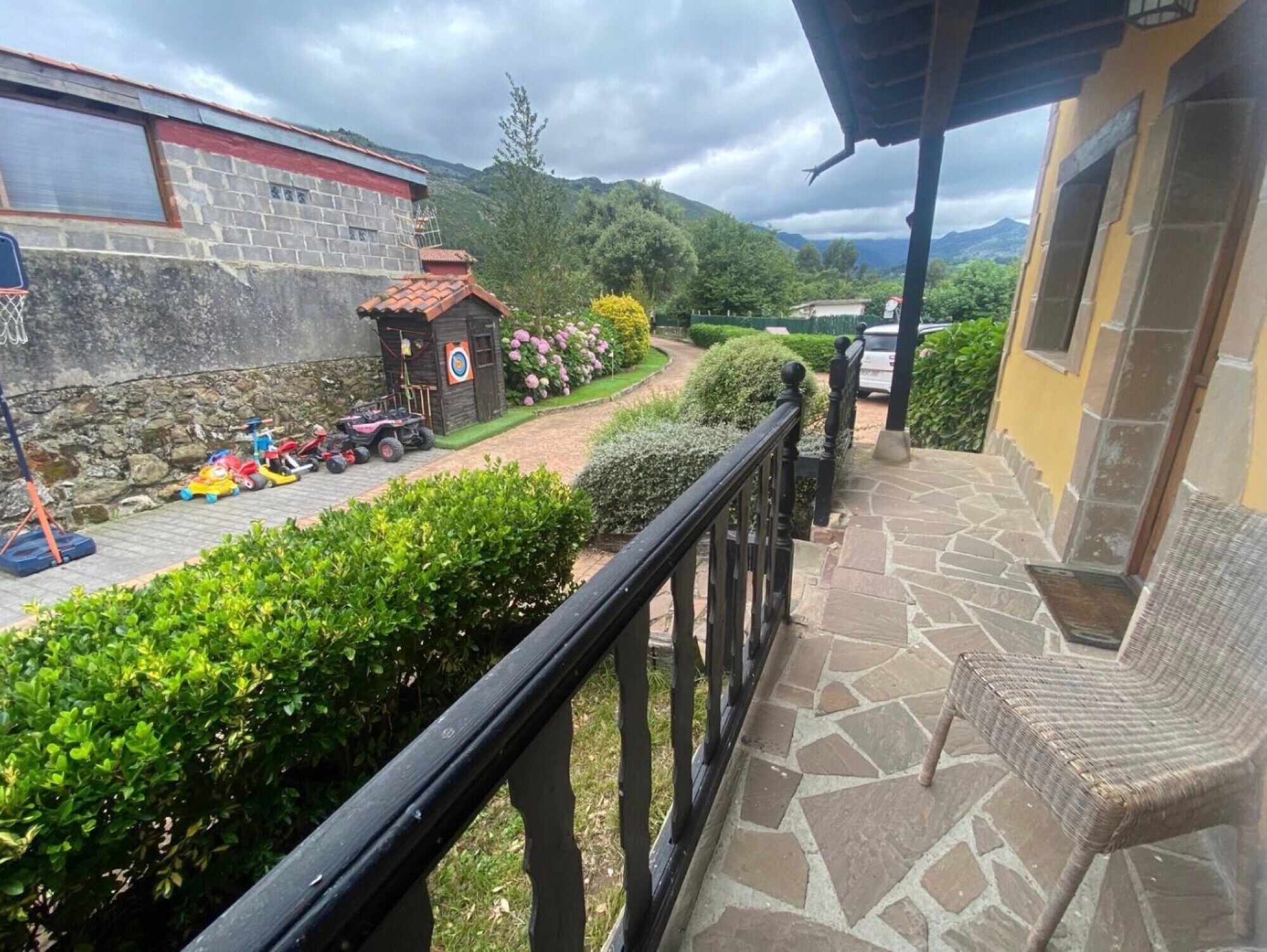 Ein Balkon mit Steinboden, einem Korbstuhl und Blick auf einen Kinderspielbereich mit Spielzeug und einem kleinen Gebäude.