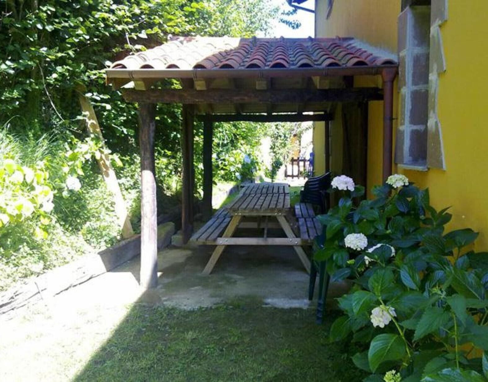 A wooden pergola with a table and chairs, surrounded by greenery and a yellow wall.