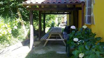 A wooden pergola with a table and chairs, surrounded by greenery and a yellow wall.