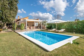 A swimming pool with a white deck, a covered patio area, and a view of a house with a tiled roof.