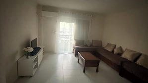 A living room with a sofa, a coffee table, a TV stand, and a window with curtains. - HOTEL NASSMA residence  (Jijel)
