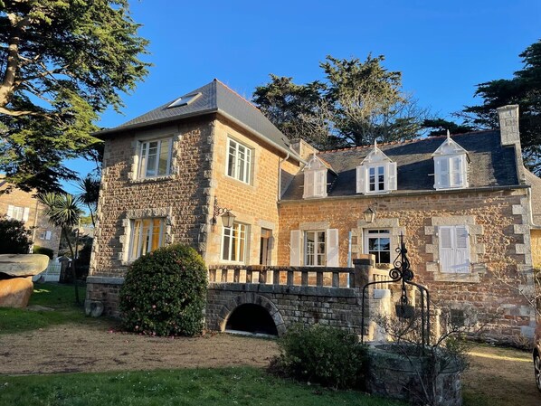 Un bâtiment en pierre avec une façade en briques, de nombreuses fenêtres dotées de volets et un pont en pierre menant à l’entrée.