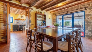 A rustic dining area with a large wooden table, chairs, and a kitchen in the background.