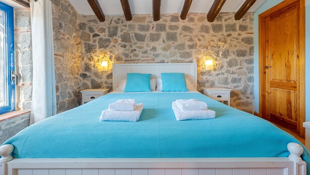 A bedroom with a stone wall, wooden beams, a bed with blue bedding, two bedside tables with lamps, and a wooden door.