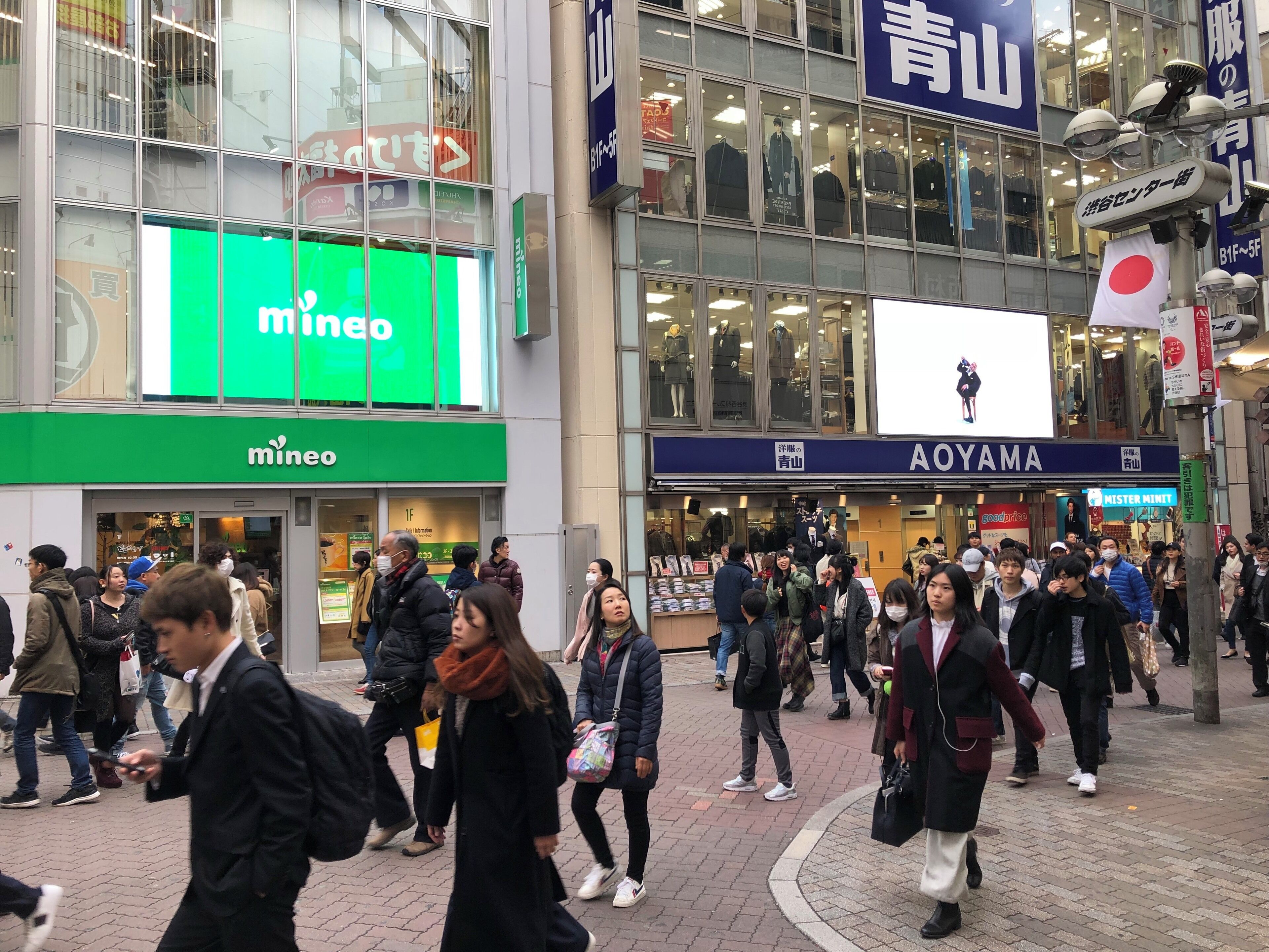 Una zona comercial urbana animada amb botigues com 'mineo' i 'Aoyama'.