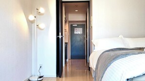 Una habitación de hotel con cama, mesita de noche, lámpara y puerta que da a otra estancia.