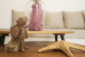 Una estatua de Buda sobre una mesa de madera, con una estrella de mar a un lado, en una habitación que tiene un sofá y un florero de vidrio al fondo.