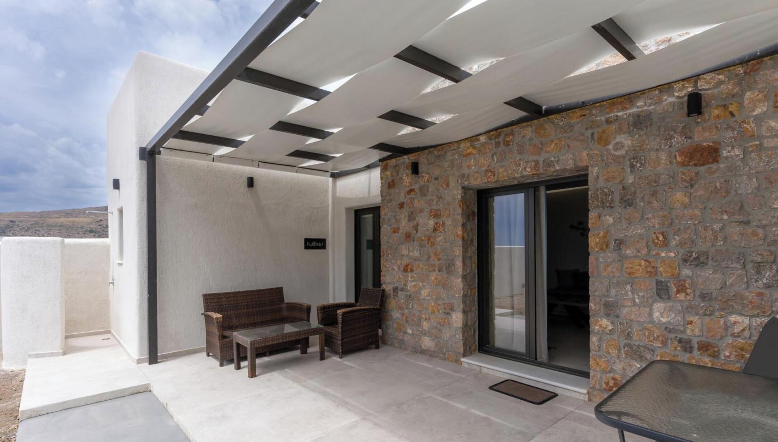 A modern outdoor patio with a stone wall, a glass door, and a seating area with a bench and chairs.
