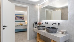 A modern bathroom with a large mirror, two stone sinks, and a bed visible through an open door.