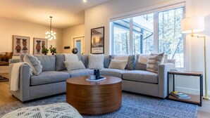 A modern living room with a sectional sofa, a round coffee table, and a large window offering a view of trees.