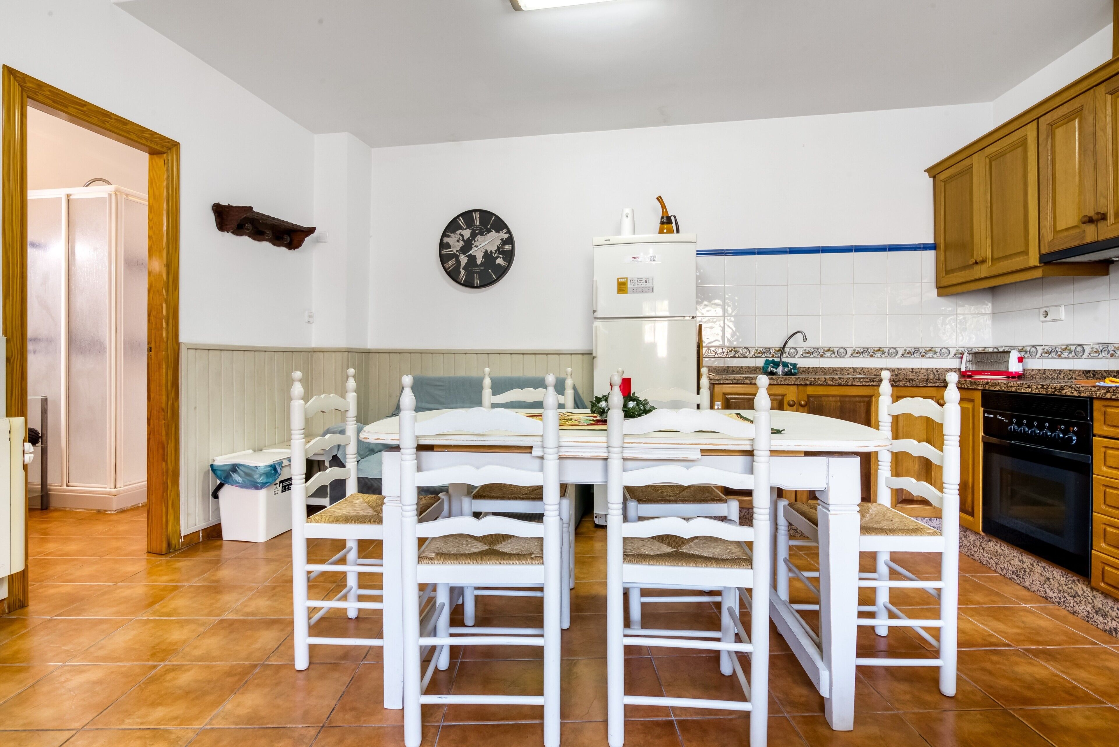 Een keuken met witte kastjes, een witte eettafel met vier stoelen, een klok aan de muur en een koelkast.