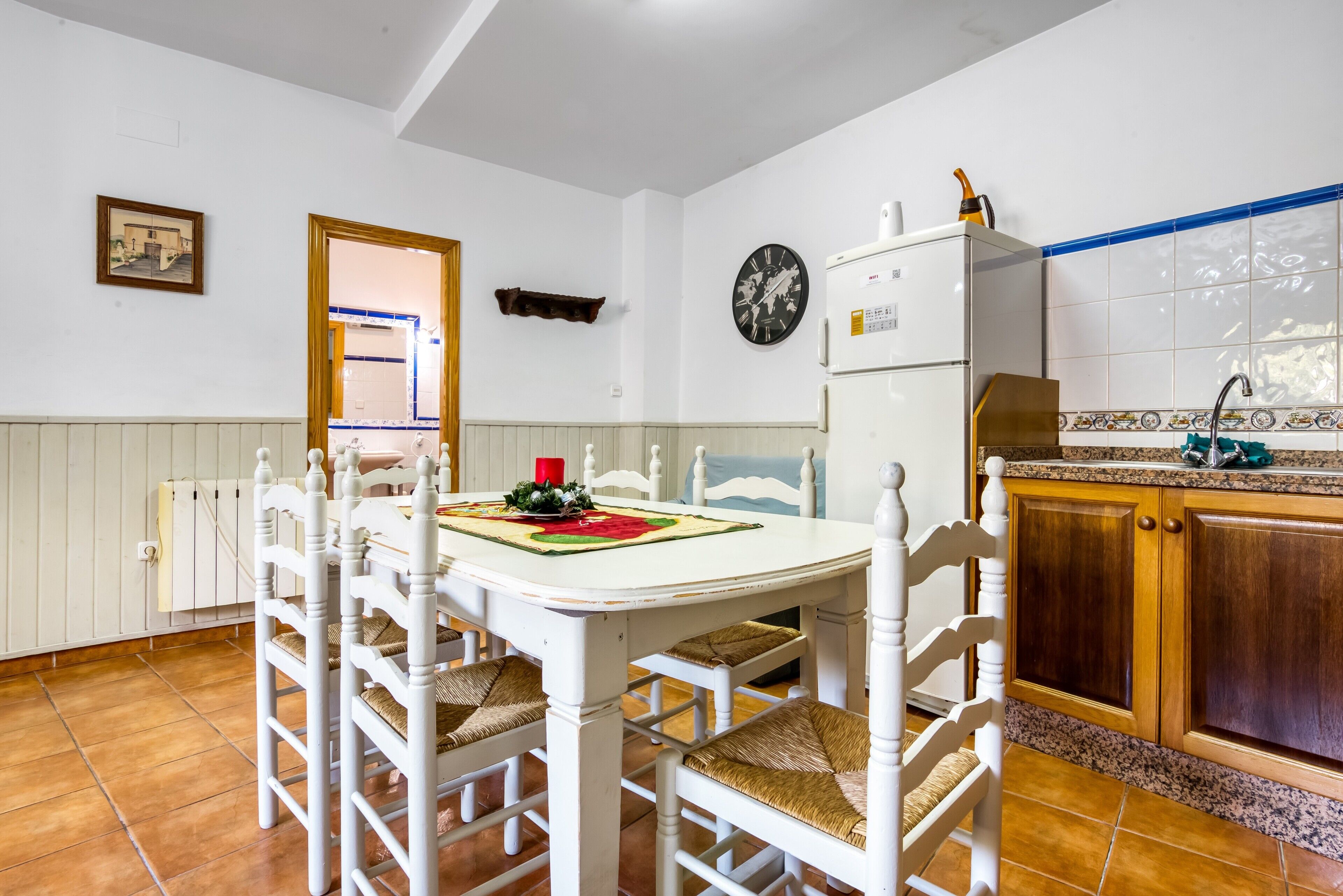 Un coin repas avec une table et des chaises blanches, une horloge au mur, et une cuisine dotée de meubles en bois et d’un évier.