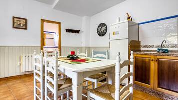 Un coin repas avec une table et des chaises blanches, une horloge au mur, et une cuisine dotée de meubles en bois et d’un évier.
