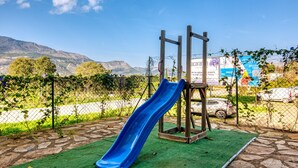 Une aire de jeux avec un toboggan bleu et une structure d’escalade, entourée d’une clôture, et des montagnes en arrière-plan.