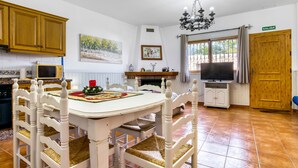 Une cuisine avec des meubles de rangement en bois, une table à manger avec des chaises, un téléviseur et un lustre.