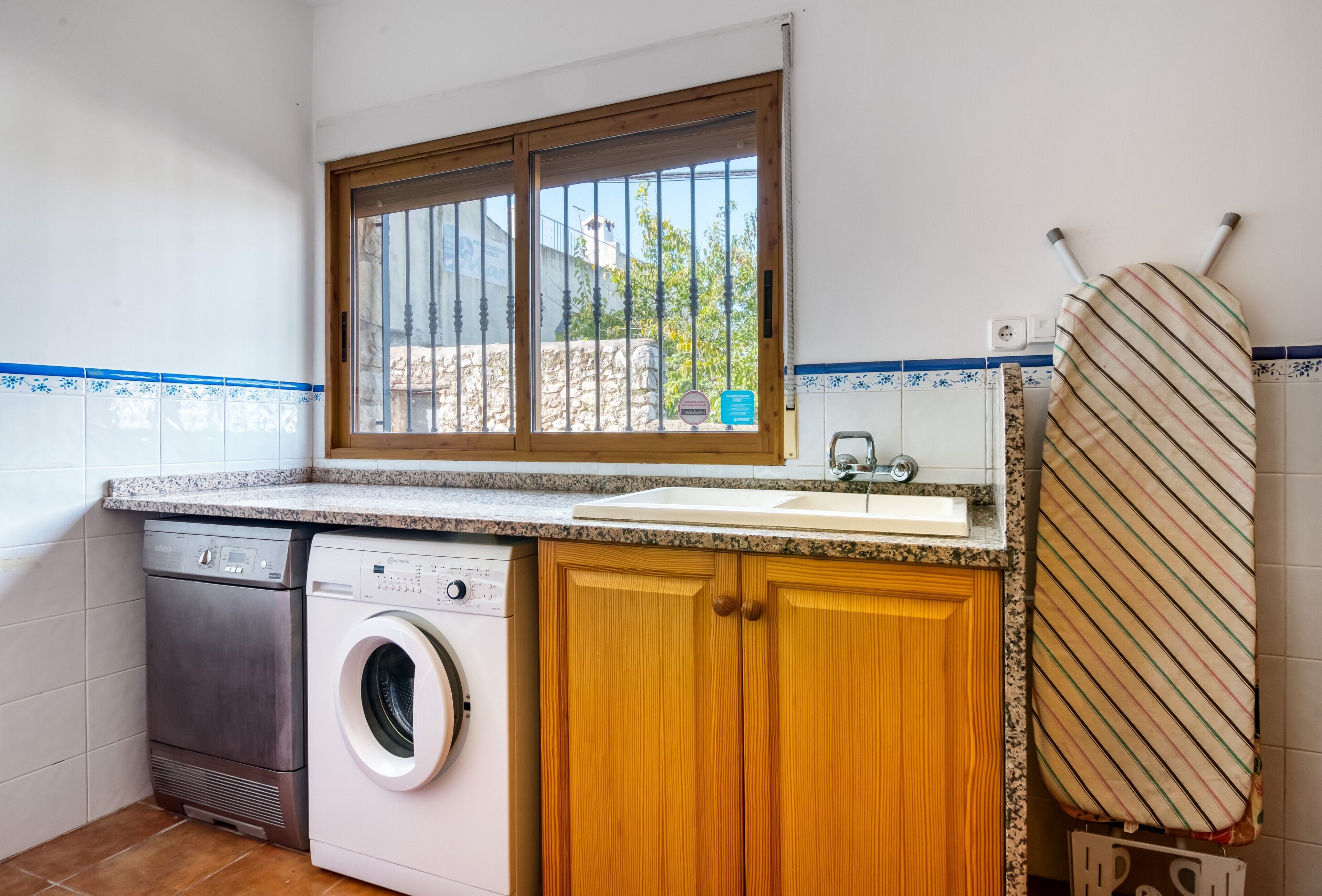 Une cuisine compacte équipée d’un lave-linge, d’un évier, de meubles en bois et d’une fenêtre donnant sur un paysage agréable.