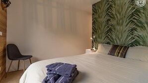 Een netjes opgemaakt bed met handdoeken, een stoel, een nachtkastje met lamp en een grote plant op de achtergrond.