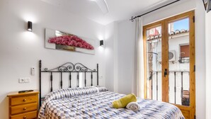 Une chambre à coucher avec une couvre-lit à motifs, une table de chevet en bois, un luminaire fixé au mur et une porte-fenêtre donnant sur un balcon.