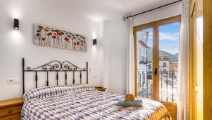 Een slaapkamer met een bed, een aan de muur hangend schilderij, een raam met gordijnen en uitzicht op gebouwen.