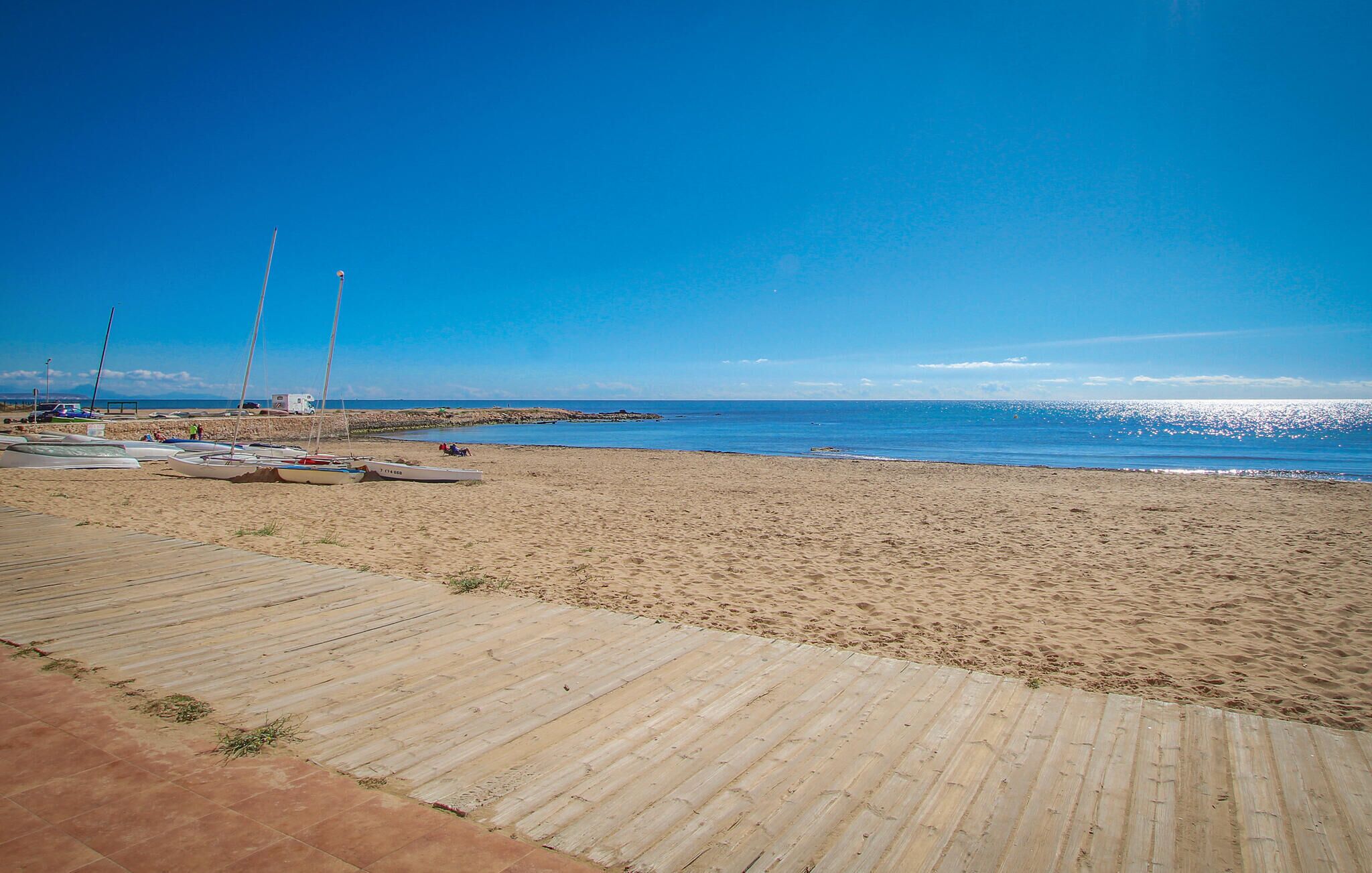 A sandy beach with a wooden walkway leading to the water, several boats, and a rocky breakwater in the distance.
