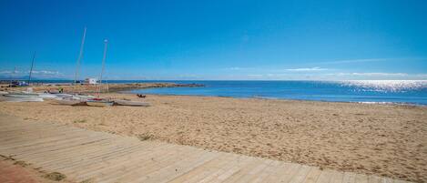A sandy beach with a wooden walkway leading to the water, several boats, and a rocky breakwater in the distance.