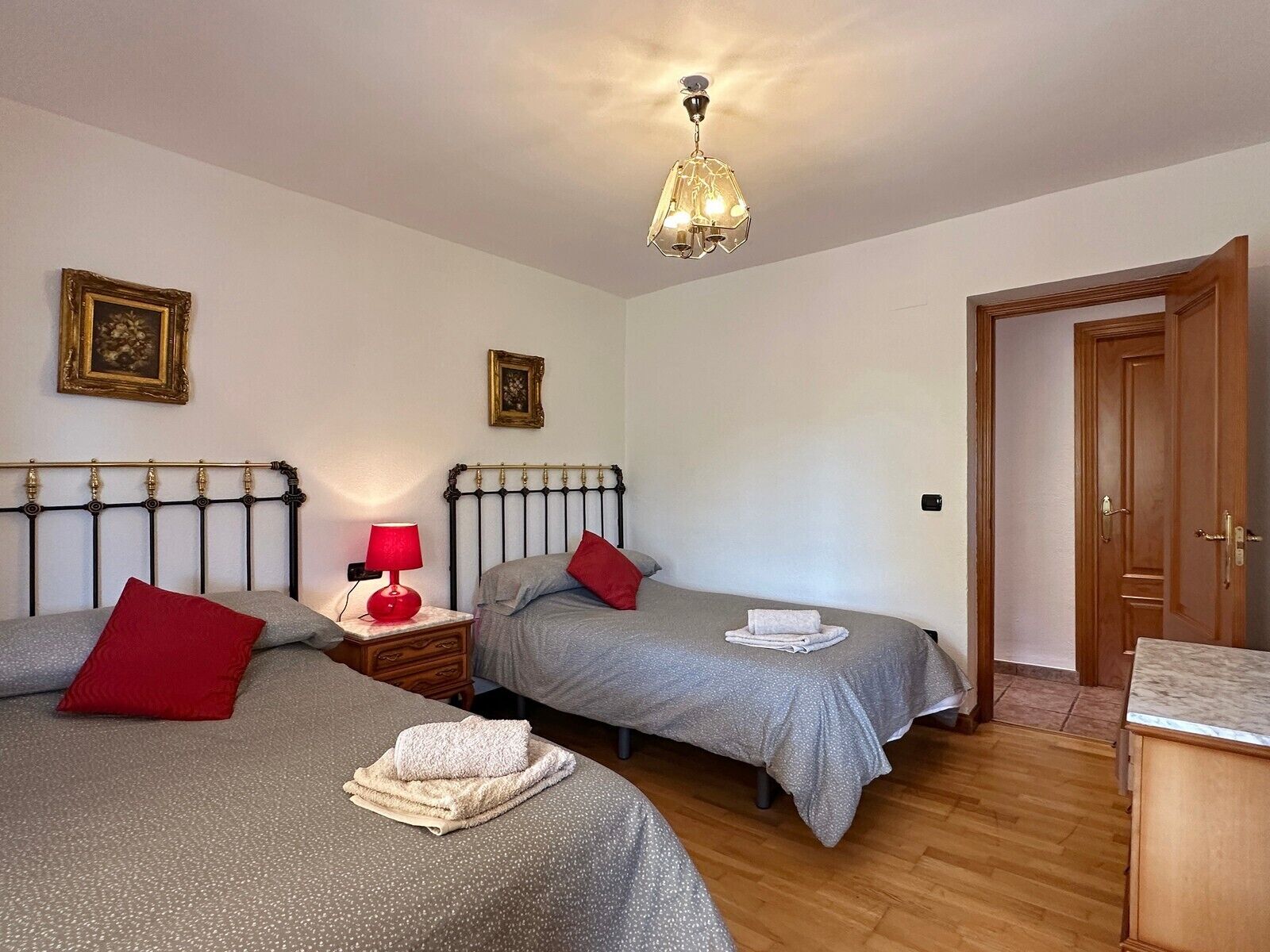 A bedroom with two beds, a nightstand with a lamp, and a door leading to another room.