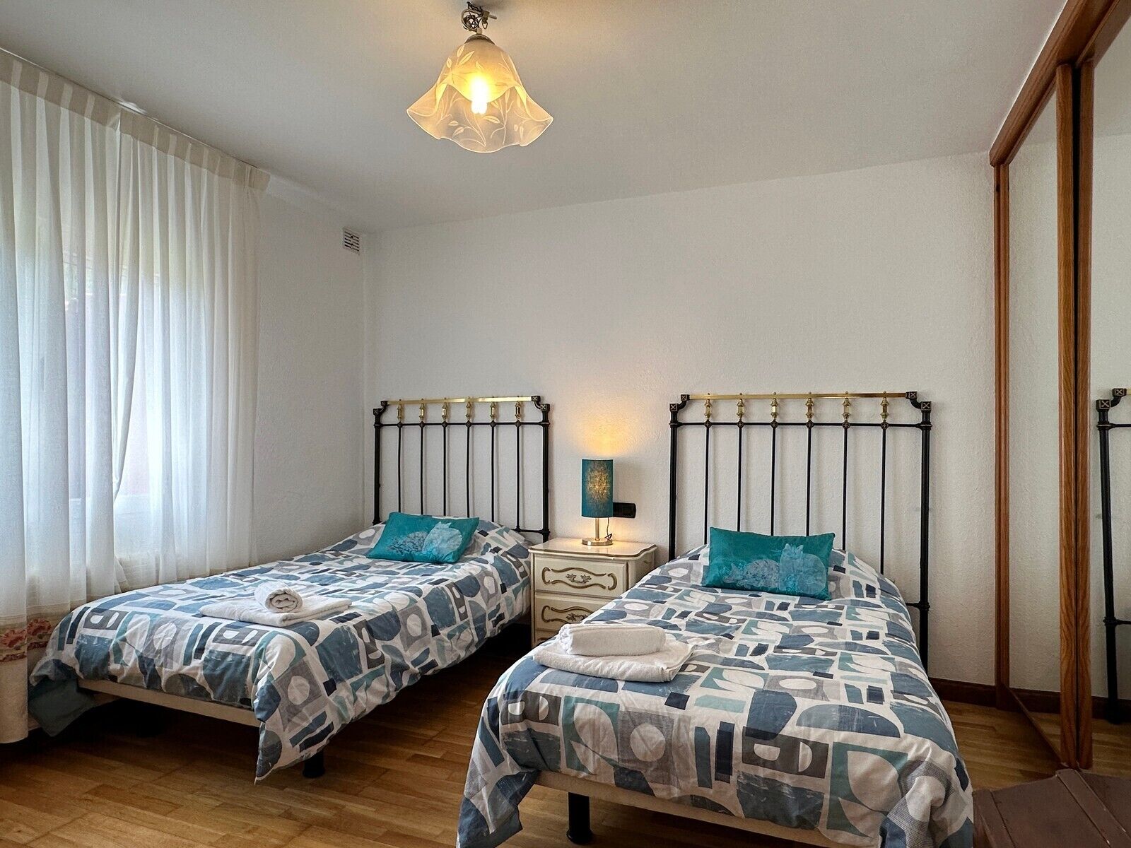 Un dormitorio con dos camas, una mesita de noche, una lámpara y una ventana con cortinas.