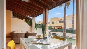 Een overdekt terras met een tafel, stoelen, een rieten bank en uitzicht op een woonwijk.