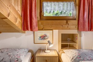 A wooden cabin room with two beds, a nightstand with a lamp, a window with curtains, a wall-mounted picture, and a shelf.